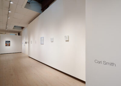 A bright art gallery with light wood floors and white walls displays several small paintings in a row. The name "Carl Smith" is written on the wall to the right. Recessed lighting illuminates the space.