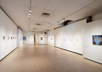 A spacious, well-lit art gallery with white walls and light wood floors displays small paintings evenly spaced along the walls. Track lighting highlights the artwork, and the room appears calm and empty.