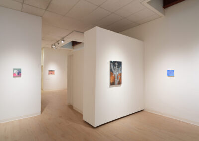 A modern art gallery with light wood floors and white walls displays several small, colorful paintings, each hung on separate wall panels under ceiling spotlights.
