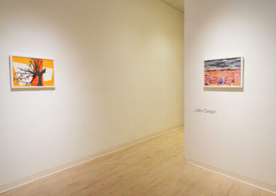 A minimalist art gallery with two colorful landscape paintings hanging on white walls. The name "Julie Green" is displayed on the wall beneath the painting on the right. The floor is light wood.