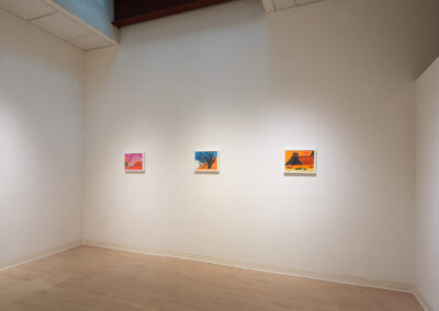 A white-walled art gallery displays five colorful abstract paintings in a row on one wall. The artworks are spaced evenly, and the floor is light wood. The name "Julie Green" appears faintly on the left wall.