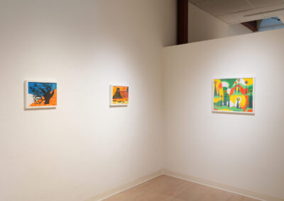 A white gallery wall displays three colorful framed paintings with abstract and figurative designs, spaced evenly on the wall in a well-lit corner.