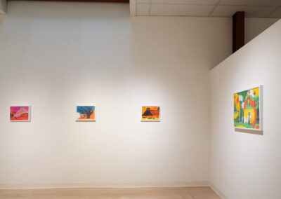 A minimalist art gallery wall displays four colorful framed paintings, evenly spaced. The first three are small and square, while the fourth is larger and rectangular, all featuring vibrant, abstract designs.