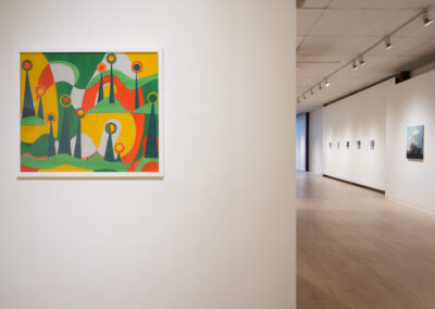 A colorful abstract painting with geometric shapes hangs on a white wall in a bright, modern art gallery. More framed artworks are displayed along the corridor in the background.