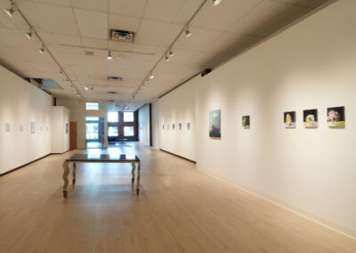 A bright, modern art gallery with white walls and light wood floors displays small paintings and drawings; a unique table with curved legs stands in the center of the room near the entrance.