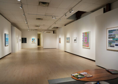 A modern art gallery with white walls displays colorful framed artworks; a wooden table with magazines sits in the foreground, and spotlights illuminate the spacious, light-wood floor interior.