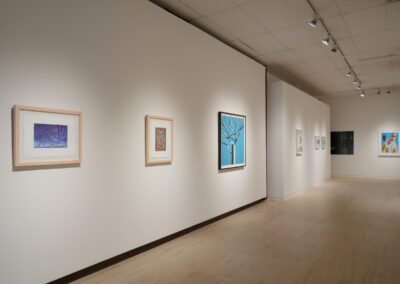 A modern art gallery with framed paintings hanging on white walls, bright overhead lighting, and a light wooden floor. The artworks vary in style and color. The space appears quiet and spacious.