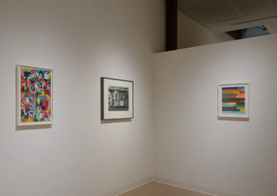 Three framed artworks hang on a white gallery wall: a colorful geometric piece on the left, a grayscale architectural photograph in the center, and a minimalist abstract with horizontal color blocks on the right.