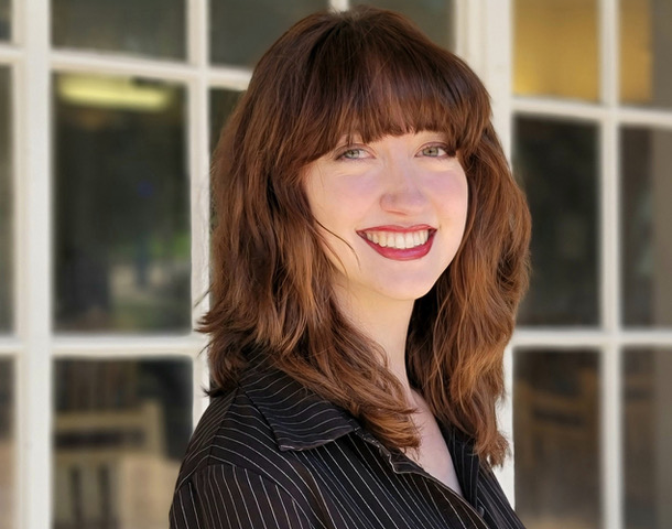 Savannah Phelps headshot- A woman with wavy brown hair and bangs smiling, wearing a black pinstripe shirt, stands in front of a window with white frames.