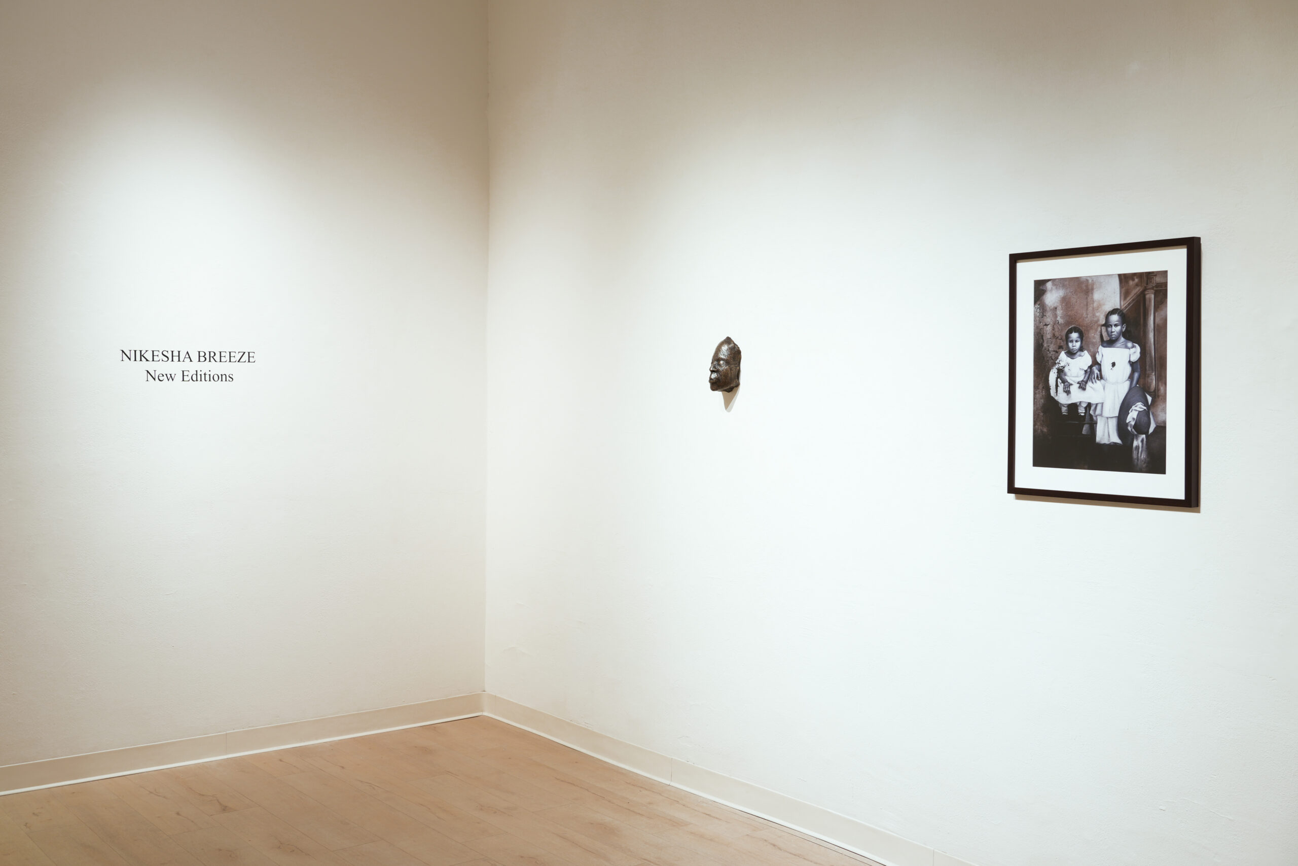 A minimalist gallery corner displays a wall label reading “Nikesha Breeze New Editions,” a small mask sculpture, and a framed black-and-white photograph of several people. The floor is light wood, and the walls are white.