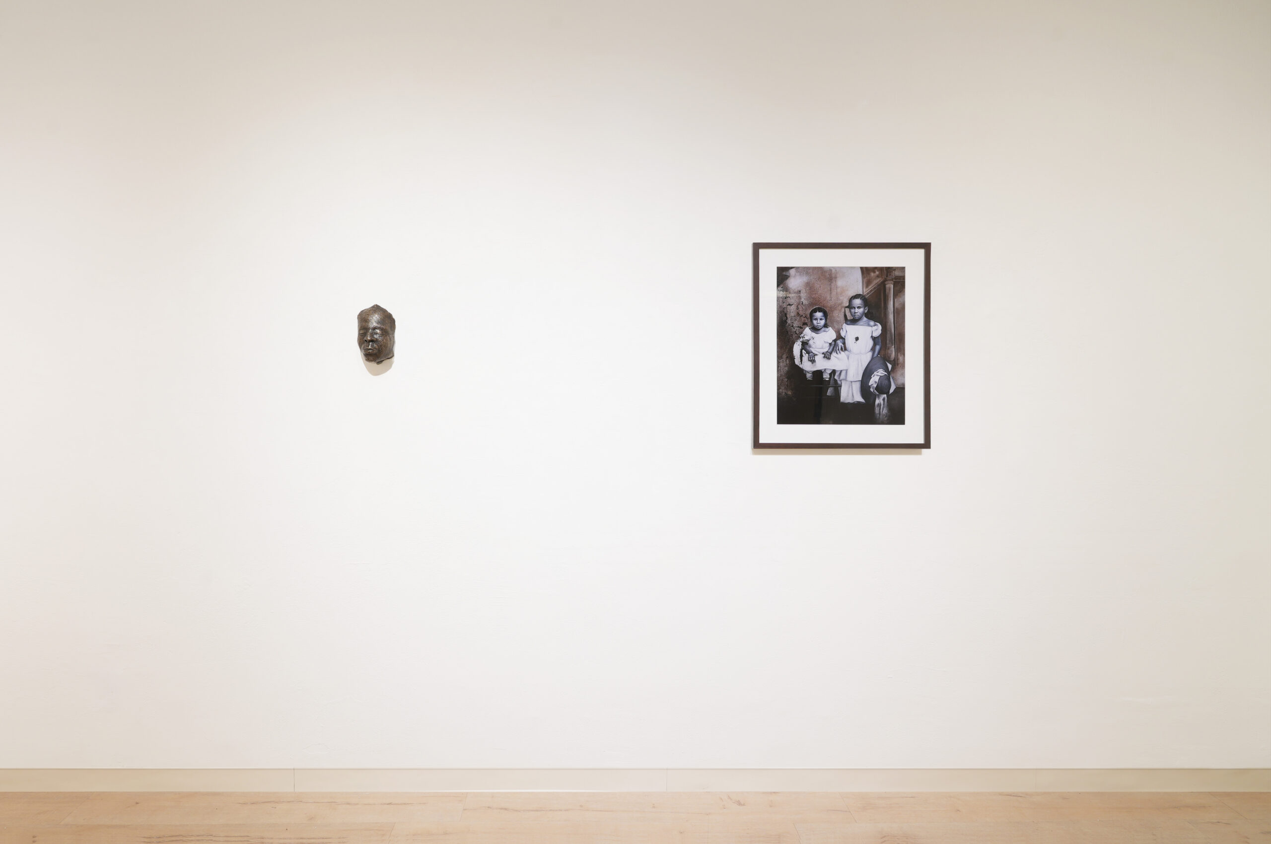 A minimal gallery wall displays a small sculpted mask on the left and a framed black-and-white photograph of three people on the right, both mounted on a plain white background above a wooden floor.