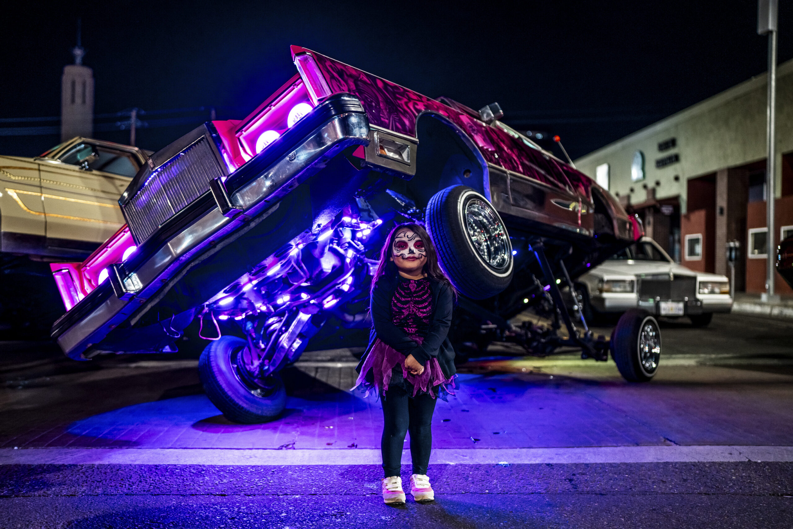 A young girl with sugar skull face paint stands in front of a lowrider car with its front end lifted high, illuminated by purple underglow lights in this Seventh and Central piece by Nathaniel Paolinelli.